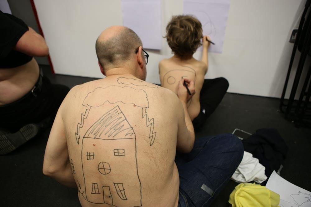 A shirtless man with a drawing of a house on his back draws on a child's back. The child draws on a paper on the wall. Playing Up Test Day.