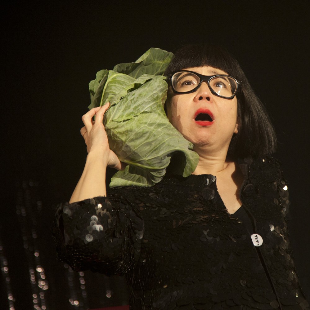 Stacy Makishi in front of a black background, holding a lettuce head next to her head