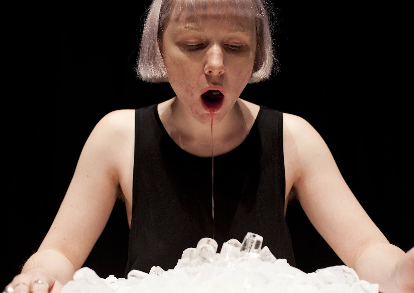 MA Live Art student Molly Honer in front of a black background. A liquid is dropping out of her mouth onto a stack of ice cubes.
