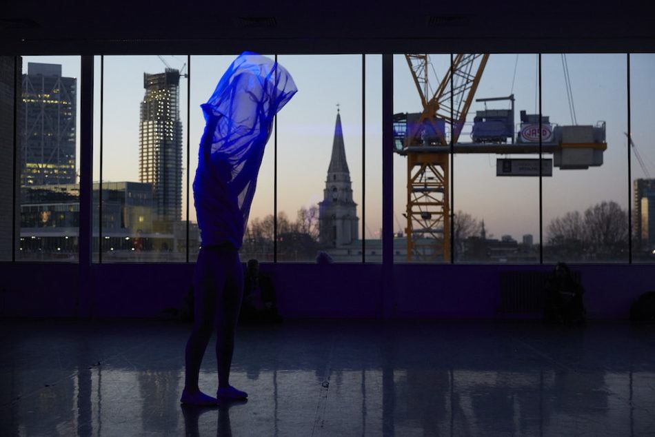 Semi nude artist holds a translucent bluwe fabric over their head, in a room overlooking the London skyline at dusk.