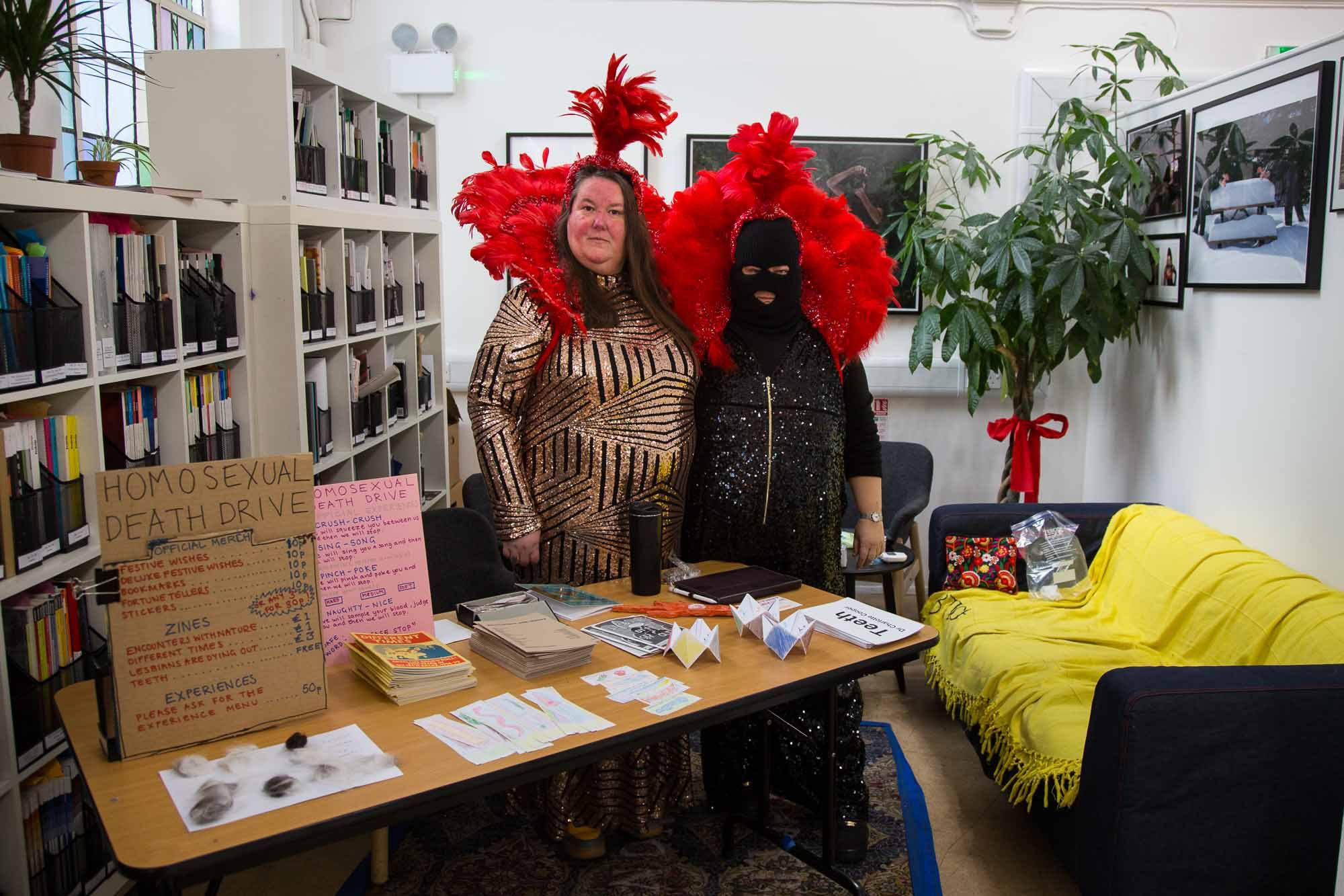 Homosexual Death Drive, a queer punk band stand behind a table of merch. They wear sparkly jumpsuits and red showgirl head pieces.