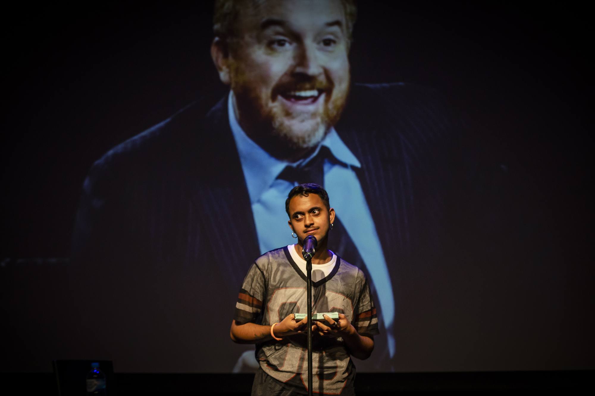 The artist speaks into a microphone as though doing a comedy set in front of an image of Louis CK.