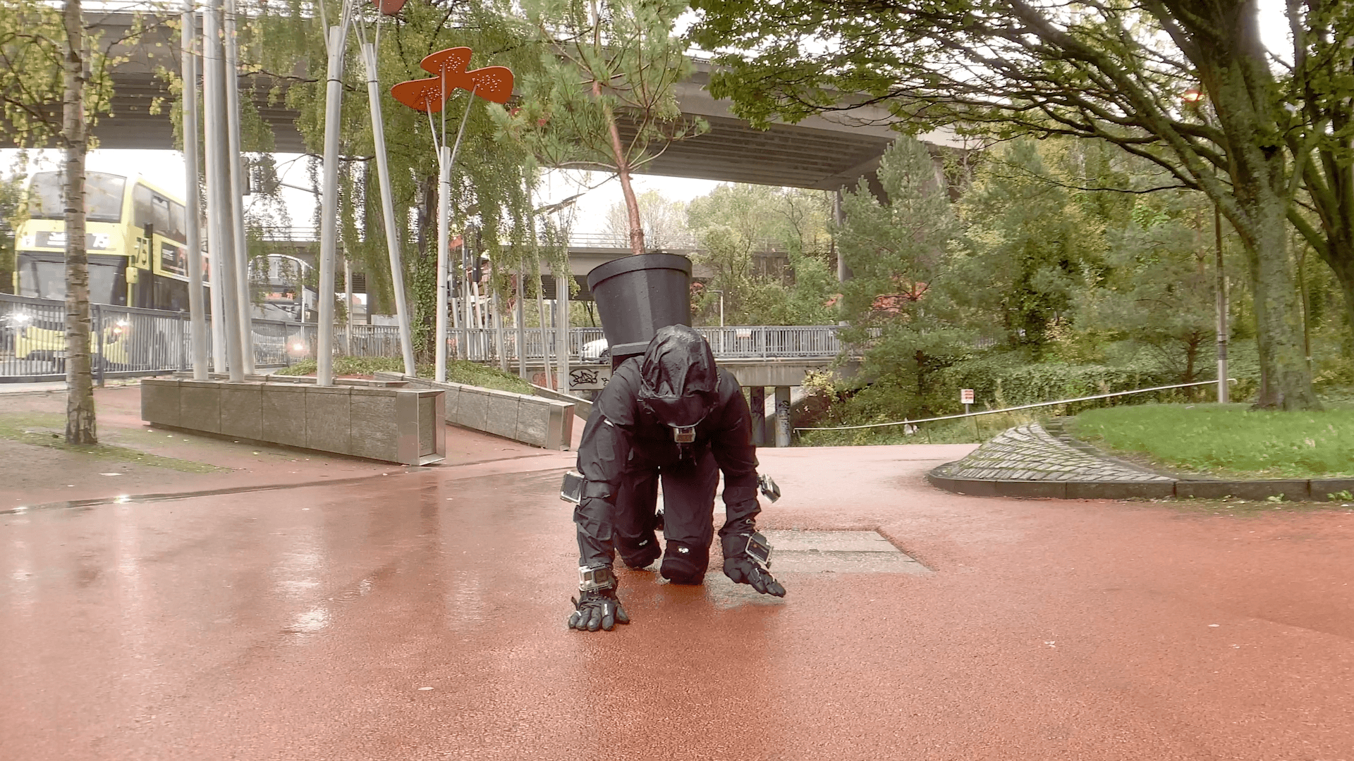A woman crawling towards the camera on her hands and knees. One of her hands is in mid-air as she takes her next hand step. She is wearing black waterproofs, knee pads and black gloves. Her hood is up so we cannot see her face. She has GoPro cameras attached to her arms, wrists, hands, legs and there is one in her mouth. She is carrying a large black plant pot on her back, with a Scots Pine tree in it. She is crawling on a wide tarmac surface. There are 3 concrete underpasses behind her, and trees and shrubs growing on grassy banks on both sides of the tarmac pedestrian way. There are two public local busses approaching her on the road on the left side of the photo.