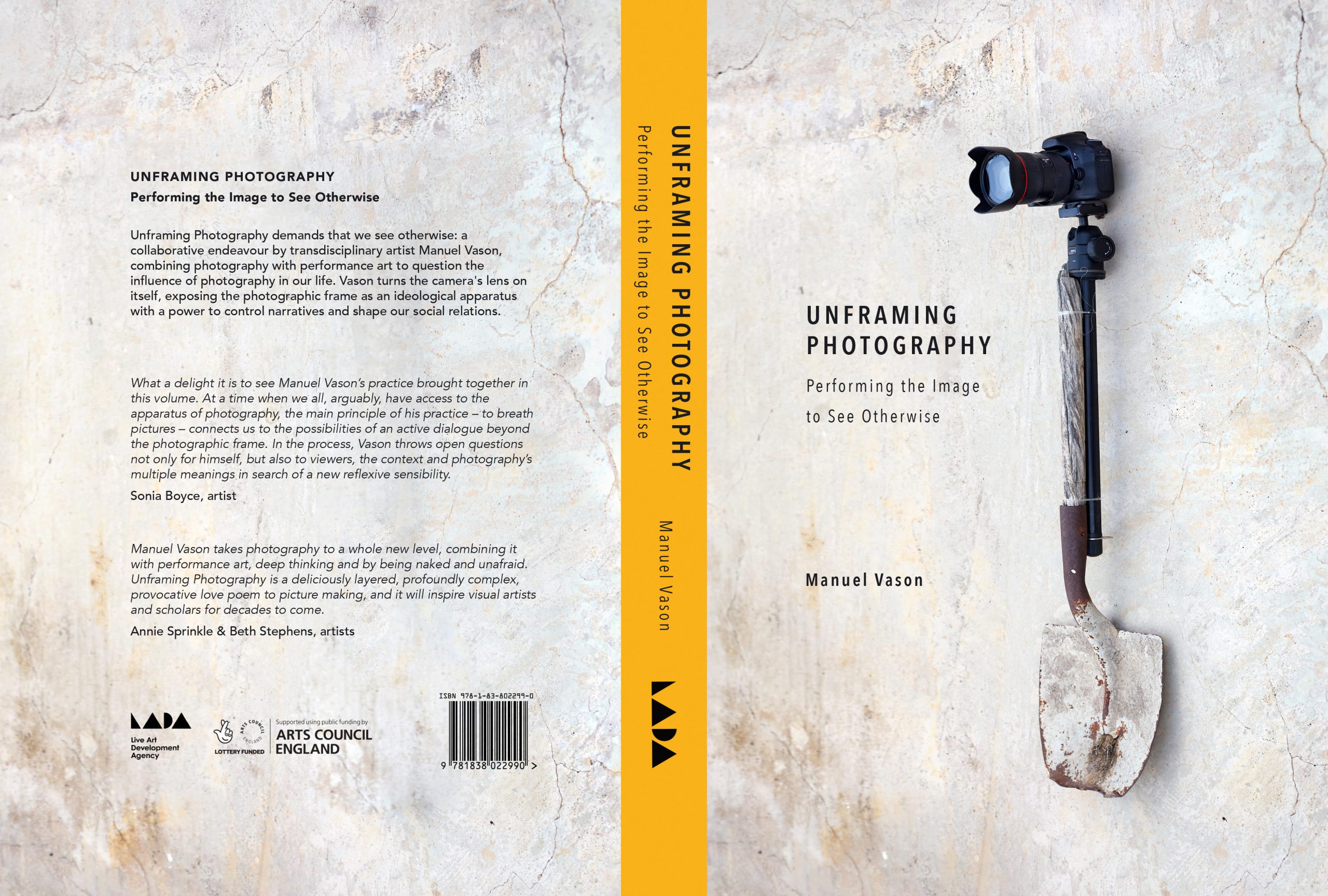 A beige concrete wall with a short shovel mounted vertically against it towards the right of the frame. The shovel is rusted and stained with concrete. The handle is wooden, of a similar colour to the concrete and is attached to a tripod and a camera with a large lens sitting on the top end. There is a thick orange line running down the left of the frame with black text adjacent reading “Unframing Photography, Performing the image to see otherwise. Manuel Vason”