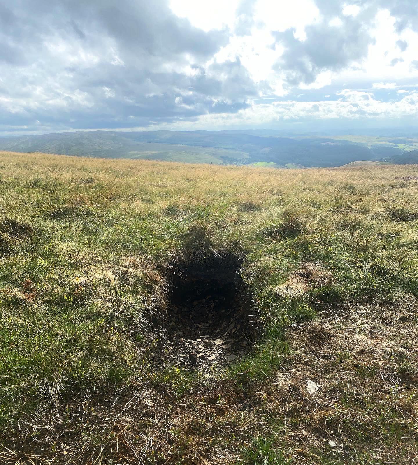 a grassy hill with a hole dug the size of a grave, looking over hilly fields on a sunny day