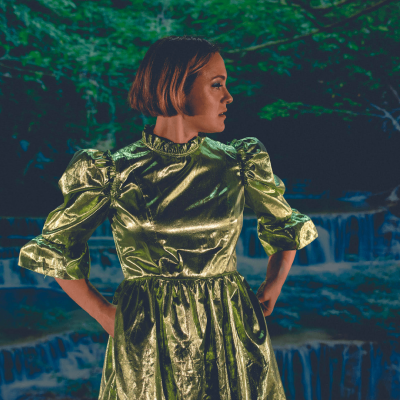 Jess Latowicki, a white woman with short brown hair with copper highlights, is stodd with her hands in the pockets of a bright green shiny dress. She is looking over her left shoulder towards a waterfall in a forest, in dusk lighting
